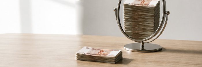 Стопка денег перед зеркалом, в отражении втрое выше — метафора разрыва между выручкой на бумаге и деньгами на счёте